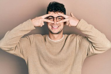 Gündelik kıyafetler giyen ve gözlük takan genç İspanyol adam barış sembolü yapıyor. Yüzünde parmaklar, gülümseyerek zaferi gösteriyor. 