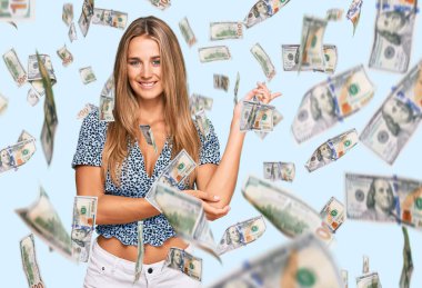 Yüzünde kocaman bir gülümseme olan sıradan kıyafetler giyen genç sarışın kadın, kameraya el ve parmak ile bakıyor..