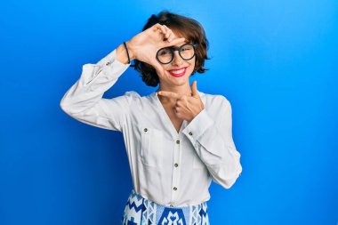 Genç esmer kadın, gündelik kıyafetler ve gözlük takıyor gülümsüyor, elleri ve parmakları gülen yüzlerle çerçeve yapıyor. yaratıcılık ve fotoğrafçılık kavramı. 