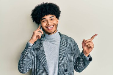 Afro saçlı genç Afrikalı Amerikalı adam akıllı telefondan konuşuyor mutlu bir gülümsemeyle el ve parmak kenarı işaret ediyor. 