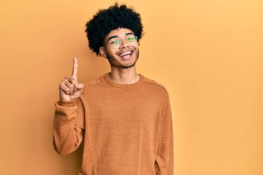 Afro saçlı, sıradan bir kazak giyen genç Afro-Amerikalı bir numarayı gösterirken kendine güvenen ve mutlu gülümseyen.. 