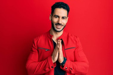 Kırmızı deri ceket giyen genç İspanyol adam elleriyle dua ediyor af diliyor kendinden emin bir şekilde gülümsüyor.. 