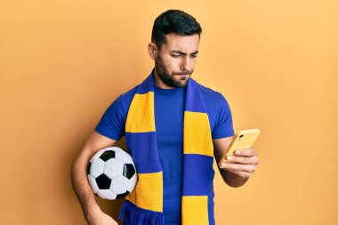 Genç İspanyol futbolcu holigan elinde futbol topu tutuyor. Akıllı telefon kuşkucu ve gergin kullanıyor. Sorunları yüzünden surat asıyor. negatif kişi. 