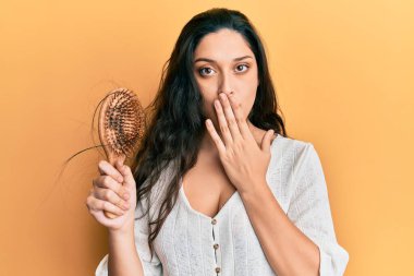 Beautiful middle eastern woman holding comb loosing hair covering mouth with hand, shocked and afraid for mistake. surprised expression  clipart
