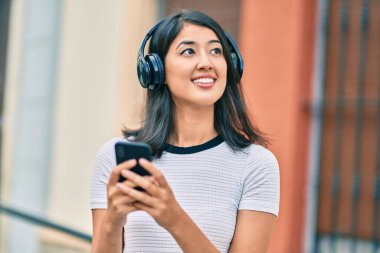 Genç İspanyol kadın şehirde akıllı telefon ve kulaklık kullanarak mutlu bir şekilde gülümsüyor..