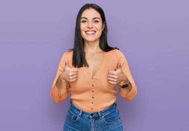 Gündelik kıyafetler giyen genç İspanyol kadın. Başarı işareti ile el hareketi yapıyor. Gülümsüyor ve mutlu. Neşeli ifade ve kazanan jest. 