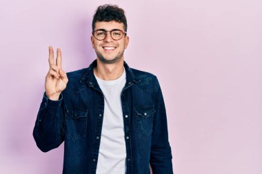 Gündelik kıyafetler giyen ve gözlüklü genç İspanyol adam iki numarayı gösterip işaret ederken kendinden emin ve mutlu bir şekilde gülümsüyor.. 