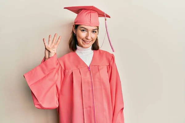 自信と幸せ笑顔ながら卒業キャップと式のローブを身に着けている若い白人女性が指の数4で現れ 指摘します — ストック写真