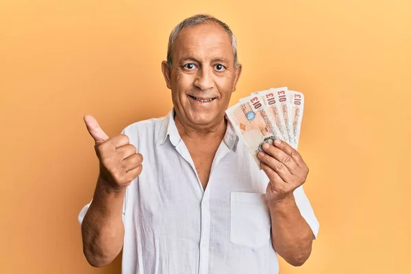 Handsome mature man holding 10 united kingdom pounds banknotes smiling happy and positive, thumb up doing excellent and approval sign