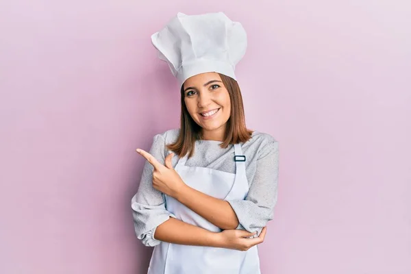 年轻美丽的女人穿着职业厨师的制服 头戴一顶愉快的帽子 满脸笑容 手指手画脚地朝旁边走去 脸上带着愉快而自然的表情 — 图库照片