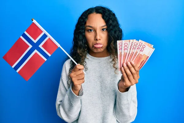 拿着诺威国旗和挪威克朗钞票的年轻拉丁女子忧心忡忡 怒气冲冲 胆战心惊 悲伤的表情 — 图库照片