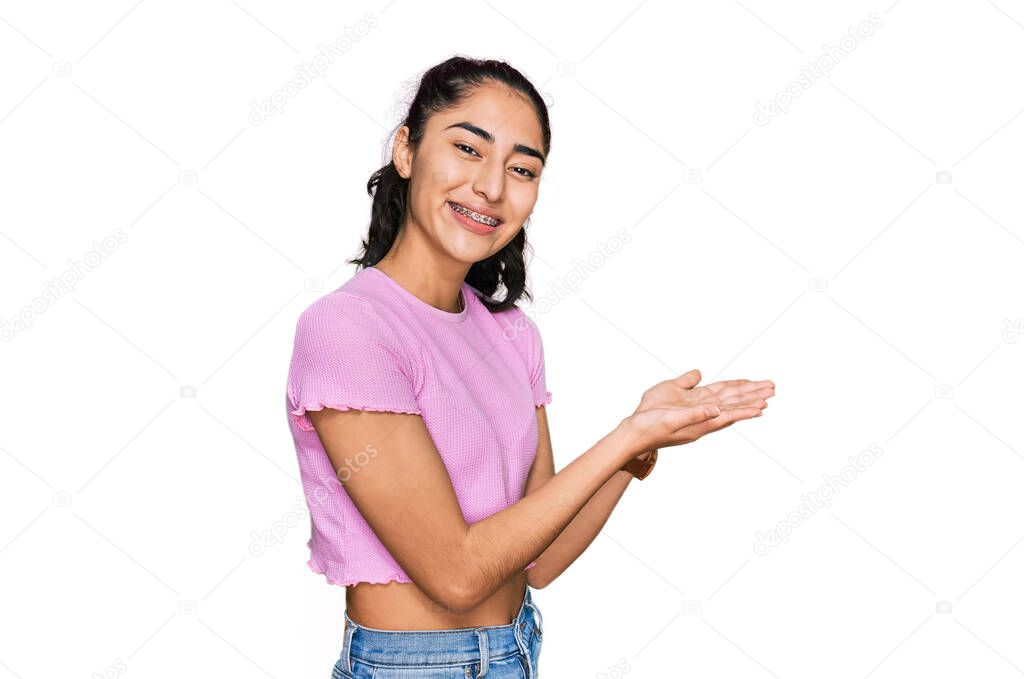 Hispanic teenager girl with dental braces wearing casual clothes pointing aside with hands open palms showing copy space, presenting advertisement smiling excited happy 