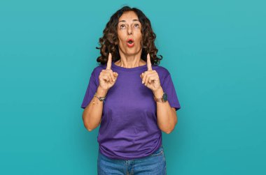 Orta yaşlı İspanyol kadın gündelik kıyafetler giyip şaşkın şaşkın yukarı bakıyor ve parmaklarıyla işaret ediyor ve kollarını kaldırıyor.. 