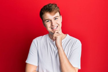 Gündelik beyaz tişört giyen genç beyaz bir adam kameraya kollarını çapraz yapıp çenesine dokunarak gülümsüyor. Olumlu düşünmek. 