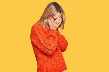Sarışın genç bir kadın, izole edilmiş sarı arka plan üzerine kışlık süveter giyiyor. Gözleri ellerle kaplıyor, hayal kırıklığına uğramış ve yorgun bir ifade.,