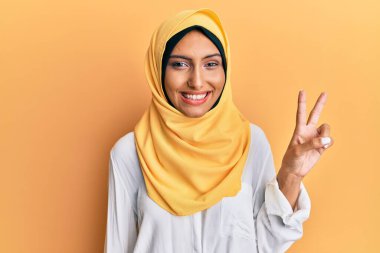 Genç esmer Arap kadın geleneksel başörtüsü takıyor. İki numarayı gösterip gösterirken, kendinden emin ve mutlu bir şekilde gülümsüyor.. 