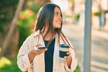 Genç İspanyol kız mutlu bir şekilde gülümsüyor. Akıllı telefon kullanıyor ve parkta kahve içiyor..