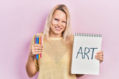 Güzel, beyaz, sarışın, elinde sanat defteri ve renkli kalemler olan bir kadın kameraya seksi bir ifadeyle bakıyor, neşeli ve mutlu bir yüzle.. 