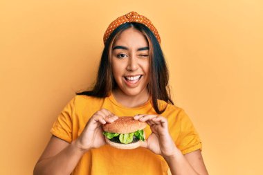 Lezzetli bir klasik burger yiyen genç Latin kadın kameraya seksi bir ifadeyle bakıyor, neşeli ve mutlu bir yüzle.. 