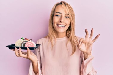 Elinde pasta tatlısı tutan güzel sarışın kadın parmaklarıyla imza atıyor, gülümseyerek mükemmel bir sembol oluşturuyor. 