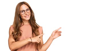 Gündelik kıyafetler giyen, gözlüklü, gülümseyen ve kameraya bakan genç İspanyol kadın iki eliyle ve parmaklarıyla kenarı işaret ediyor.. 