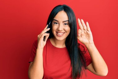 Genç, güzel, İspanyol bir kız akıllı telefonda konuşuyor parmaklarıyla imza atıyor, gülümsüyor ve mükemmel bir sembol oluşturuyor. 