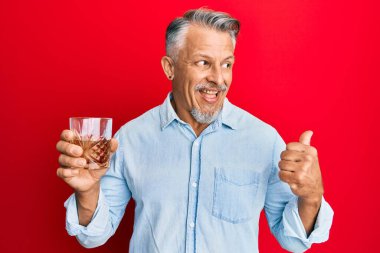 Orta yaşlı, kır saçlı, viski bardağı içen, baş parmağını kaldıran, ağzı açık gülümseyen bir adam. 