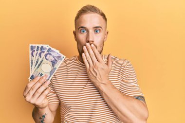 Elinde Japon yen banknotları tutan genç beyaz bir adam ağzı elle kapatıyor, şaşırmış ve hata yapmaktan korkuyor. şaşırmış ifade 
