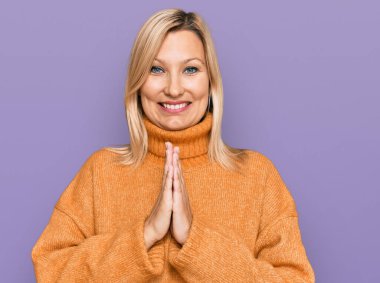 Orta yaşlı, beyaz bir kadın, sıradan bir kazak giyiyor, elleriyle dua ediyor, af diliyor, kendinden emin bir şekilde gülümsüyor.. 