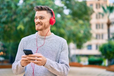 Parkta akıllı telefon ve kulaklık kullanarak gülümseyen beyaz bir genç..