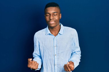 Genç Afro-Amerikan erkeği, gündelik giysiler içinde çok mutlu ve heyecanlı bir şekilde kolları havada, gülümseyerek ve başarı için çığlık atarak kazanan bir jest yapıyor. kutlama konsepti. 