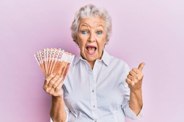 Elinde 100 Norveçli banknot tutan yaşlı, kır saçlı bir kadın başparmağıyla yan tarafı işaret ediyor ve ağzı açık gülümsüyor. 