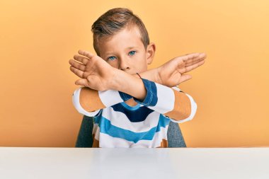 Beyaz elbiseli sevimli bir çocuk masanın üzerinde oturuyor. Reddedilmiş yüz ifadesi kollarını kavuşturuyor ve avuç içleri negatif işaretler yapıyor. Kızgın yüz ifadesi. 