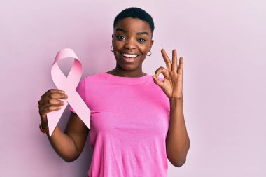 Genç Afrikalı Amerikalı kadın pembe kanser kurdelesi tutuyor parmaklarıyla imza atıyor, gülümseyerek mükemmel bir sembol oluşturuyor. 