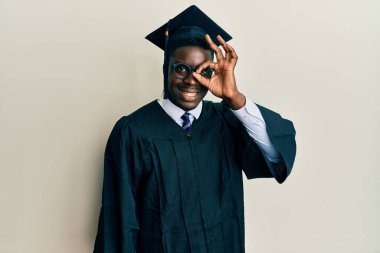 Yakışıklı siyah adam mezuniyet şapkası ve tören cübbesi takıyor. El hareketleriyle gülümsüyor, gözleriyle mutlu yüzle parmaklarının arasından bakıyor.. 