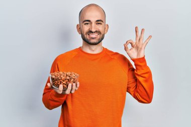 Fıstık tutan genç kel adam parmaklarıyla imza atıyor, gülümseyerek mükemmel bir sembol oluşturuyor. 