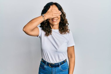 Orta yaşlı İspanyol kadın, sıradan beyaz tişörtler giyiyor, gülümsüyor ve elleri yüzünde, şaşırmak için gözleri kapatıyordu. kör kavram. 