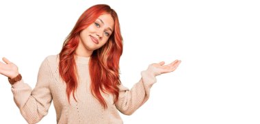 Kış süveteri giyen genç kızıl saçlı kadın, elleri ve kolları havada, kafası karışık bir ifade takınıyor. şüphe kavramı. 