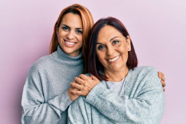 İspanyol anne ve kızı mutlu bir şekilde gülümsüyor ve soyutlanmış pembe arka planda duruyorlar..