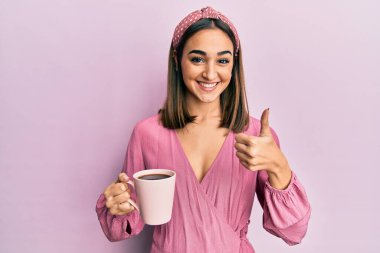 Genç esmer kız bir fincan kahve içiyor mutlu ve pozitif gülümsüyor, başparmağıyla mükemmel ve onay işareti yapıyor. 