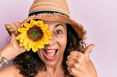 Orta yaşlı İspanyol bir kadın, yaz şapkası takıyor. Gözünün üzerinde ayçiçeği var. Parmağını kaldırıp, ağzı açık gülümsüyor. 