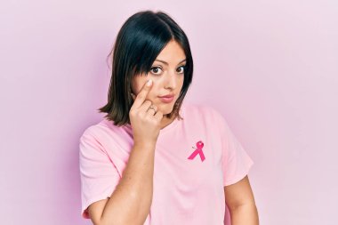 Tişörtüne pembe kanser kurdelesi takmış genç İspanyol kadın jestini izlerken gözüne işaret ediyor. Şüpheli bir ifade. 