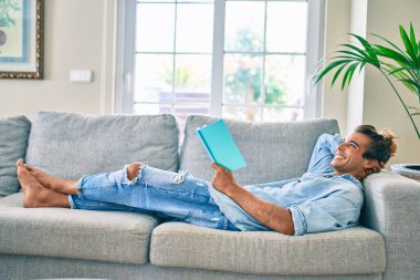Genç İspanyol gülümseyen mutlu bir kitap okuyucu evdeki kanepede uzanıyor.