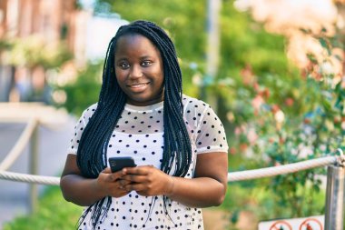 Genç Afrikalı Amerikalı kadın şehirde akıllı telefon kullanarak mutlu bir şekilde gülümsüyor..