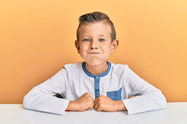 Kafkasyalı sevimli bir çocuk, gündelik giysiler giyip masada oturmuş komik suratıyla yanaklarını şişiriyor. Havayla şişmiş ağız, çılgın bir ifade.. 