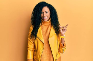 Orta yaşlı Afro-Amerikalı kadın yün kazak giyiyor ve yüzünde kocaman bir gülümseme olan deri ceket, kameraya el parmağı ile bakıyor.. 