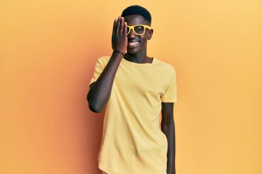 Genç Afrikalı Amerikalı bir adam günlük kıyafetler ve gözlük takıyor. Bir gözünü eliyle kapatıyor. Yüzünde kendine güvenen bir gülümseme ve sürpriz bir duygu var.. 