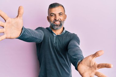 Orta yaşlı, sıradan kıyafetler giyen yakışıklı bir adam kameraya bakıp sarılmak için kollarını açmış gülümsüyor. Mutluluğu kucaklayan neşeli bir ifade.. 