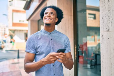 Genç Afrikalı Amerikalı adam şehirde akıllı telefon kullanarak mutlu bir şekilde gülümsüyor..