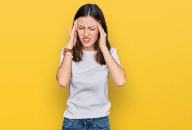 Genç, güzel bir kadın, günlük beyaz tişörtler giyiyor, elleri başının üstünde, stresten baş ağrısı çekiyor. migren ağrısı. 